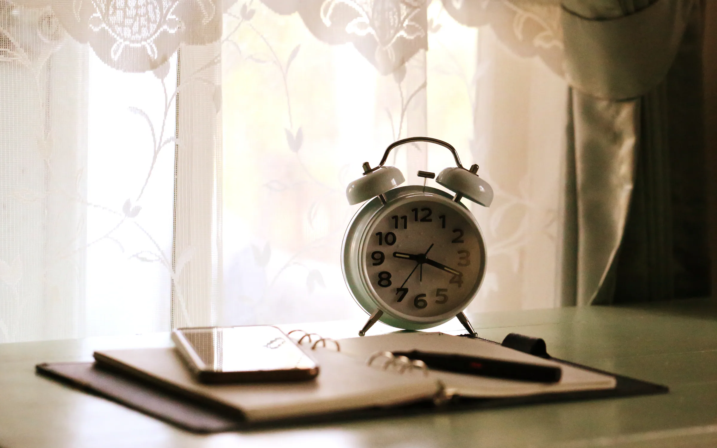 An alarm clock, a planner and a mobile phone on a table by the window
