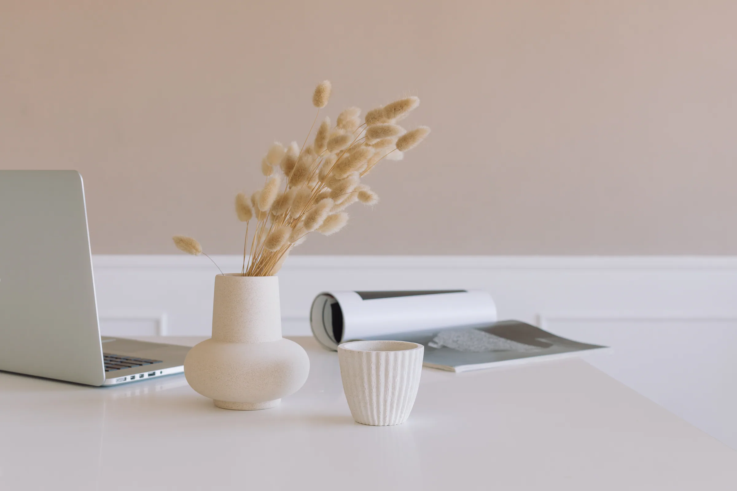 A laptop on a light desk and a vase of dried grasses